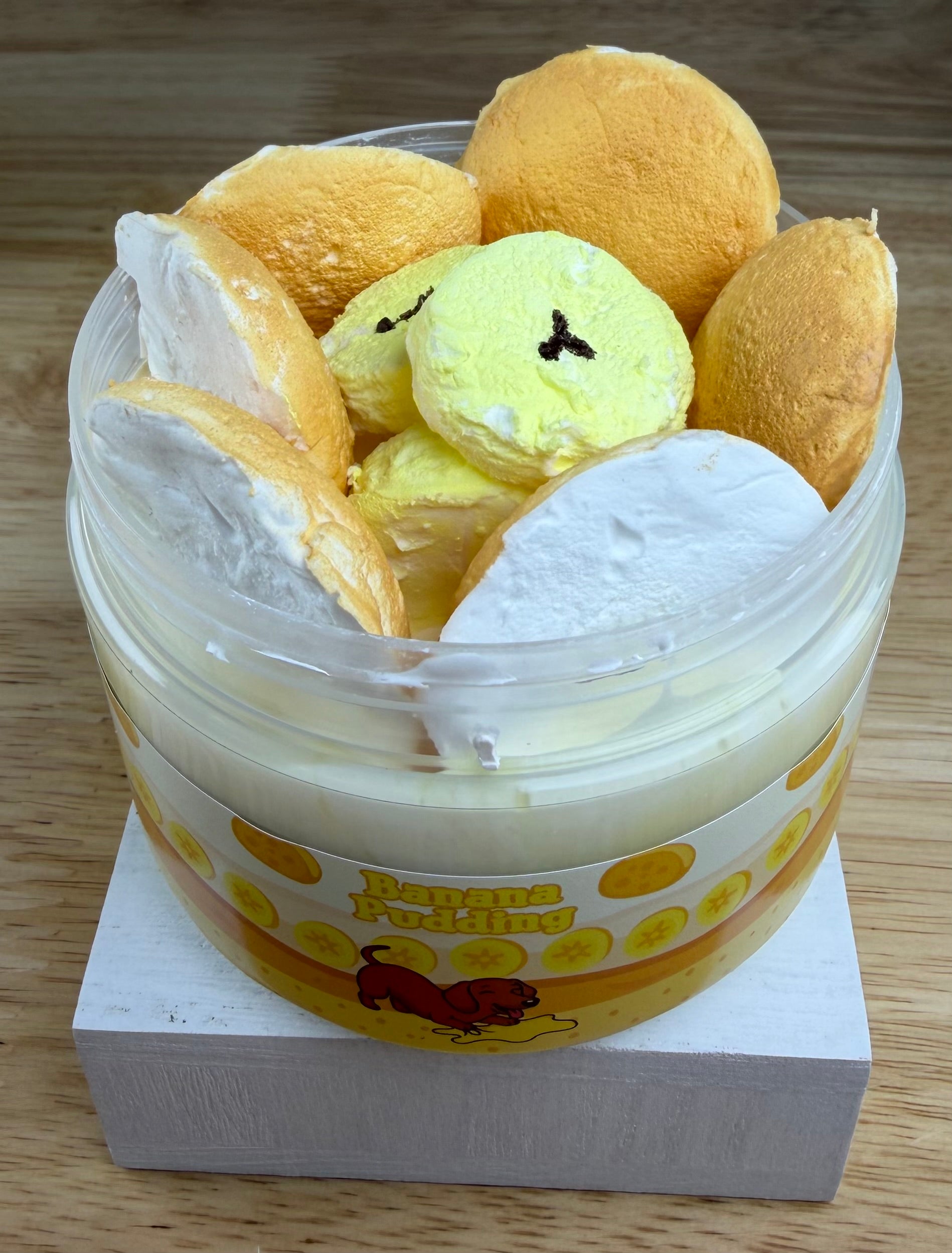 A novelty jar of 'Banana Pudding' slime, featuring a plastic jar with a yellow and orange label, topped with yellow clay banana slices with light tan clay cookie wafers, displayed on a white block.