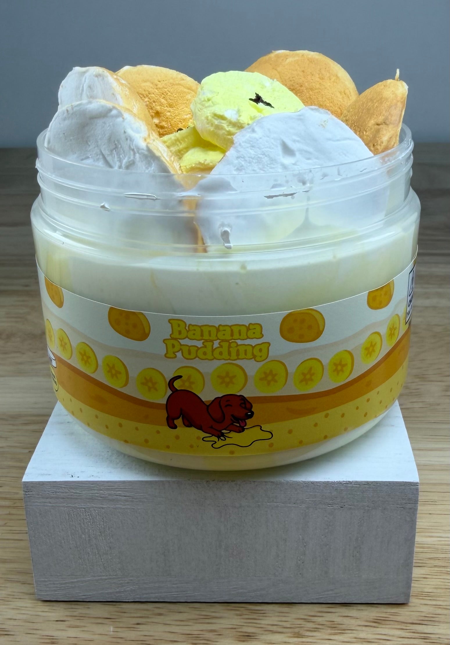 A novelty jar of 'Banana Pudding' slime, featuring a plastic jar with a yellow and orange label, topped with yellow clay banana slices with light tan clay cookie wafers, displayed on a white block.