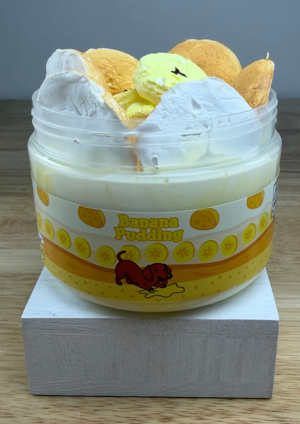 A novelty jar of 'Banana Pudding' slime, featuring a plastic jar with a yellow and orange label, topped with yellow clay banana slices with light tan clay cookie wafers, displayed on a white block.