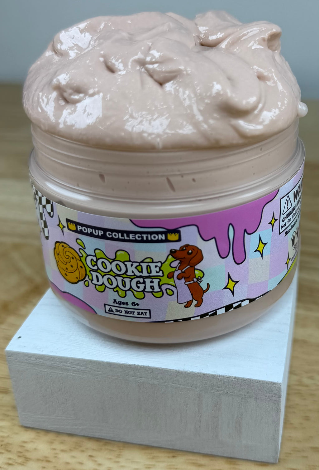 Light tan thick and glossy slime in a plastic jar labelled pop up collection with a dog and a cookie dough logo labelled cookie dough