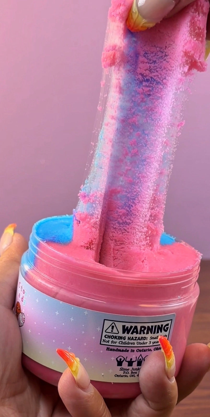 A hand holds a jar of half pink and half blue slime with a colorful pastel gradient label, stretching a long, pink and blue cloud slime strand upward against a pink background.