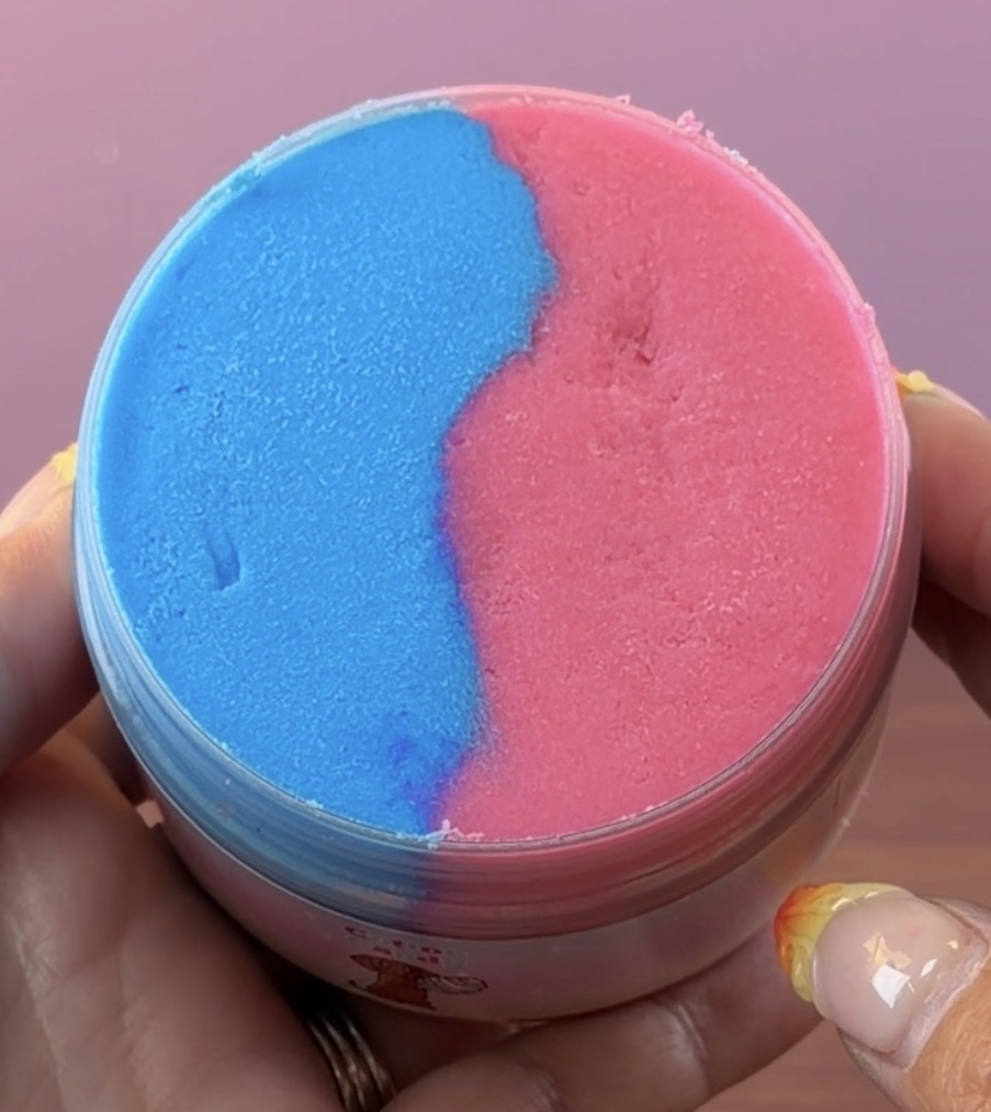 A round clear plastic jar filled with a textured blue and pink cloud slime, held in two hands. The product is split evenly into blue on the left and pink on the right, with a small cartoon graphic visible on the lower part of the jar.