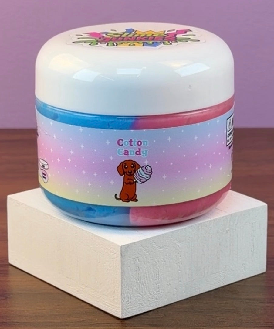 A round plastic jar of cotton candy  slime with a colorful pastel gradient label reading 'Cotton Candy' and a cartoon dachshund holding a ball of cotton candy, displayed on a white block against a purple background.