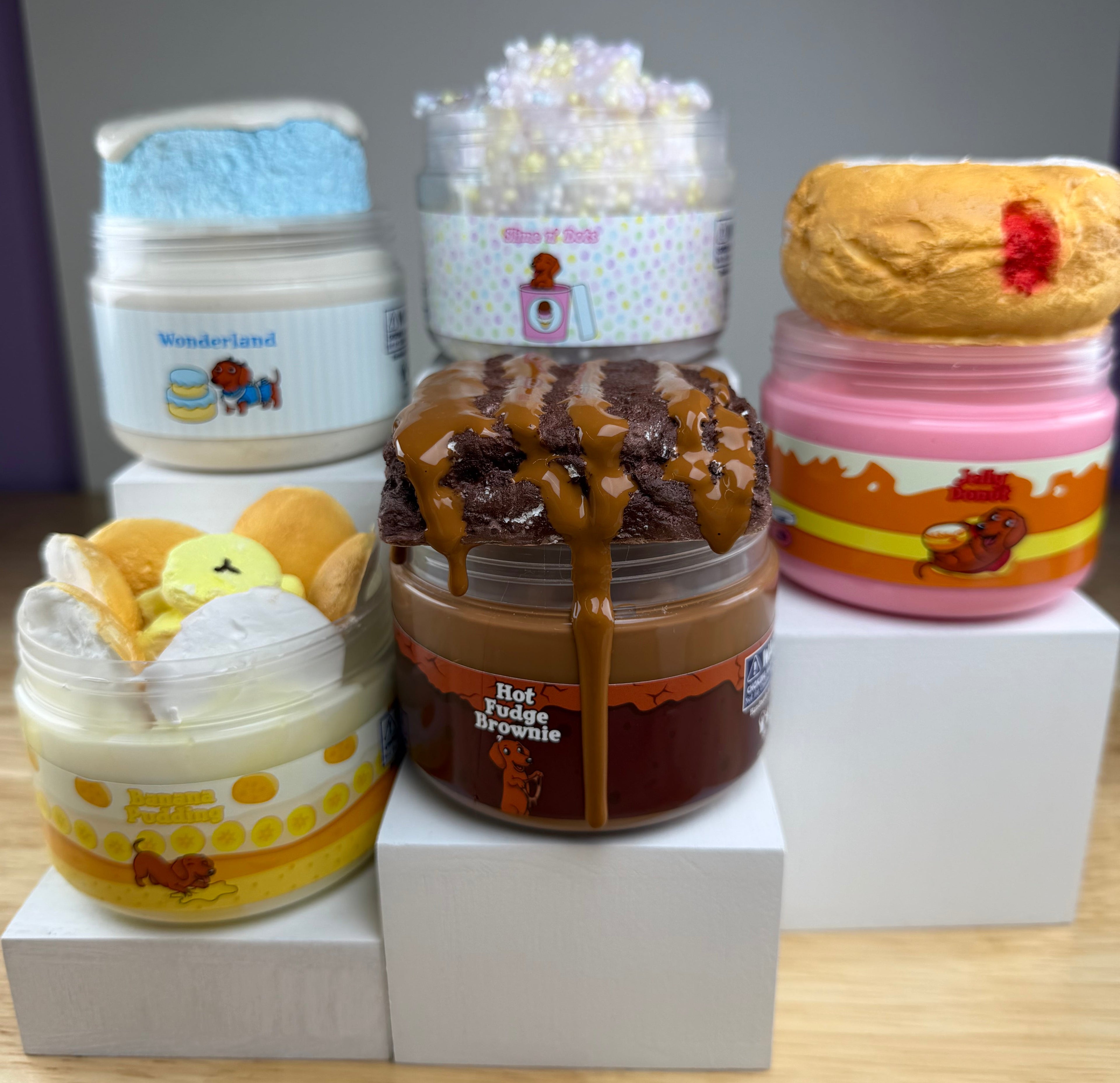 Five jars of squishy slime displayed on white display blocks on a wooden surface. The jars feature creative, food-inspired slimes with clay toppers resembling donuts, cakes, bananas, cookie wafers, and brownies with a chocolate slime drizzle.