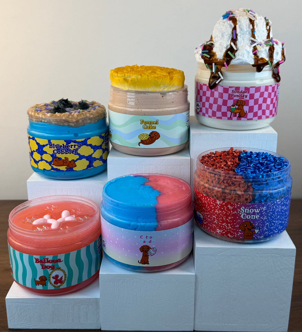 Six jars of themed slime displayed on white display blocks against a plain beige wall. The jars feature colorful cartoon-themed labels and lids, including designs resembling a blueberry cobbler, funnel cake, cotton candy, balloon dog, ice cream sundae, and snow cone.