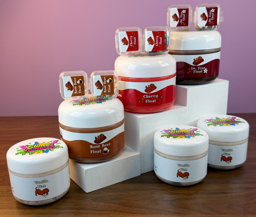 A set of slime jars with colorful labels, arranged on white display blocks over a wooden table against a pink background.