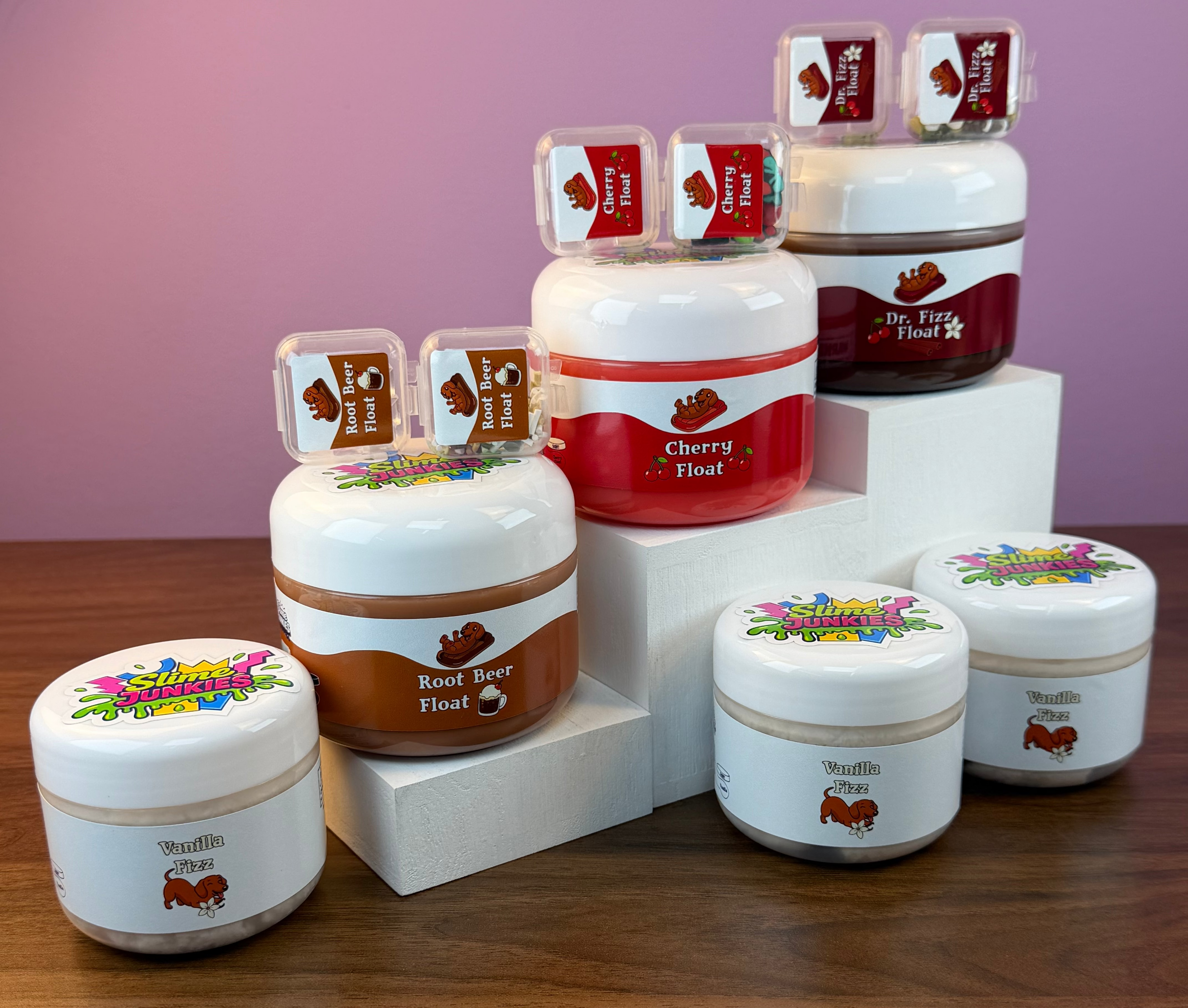 A set of slime jars with colorful labels, arranged on white display blocks over a wooden table against a pink background.