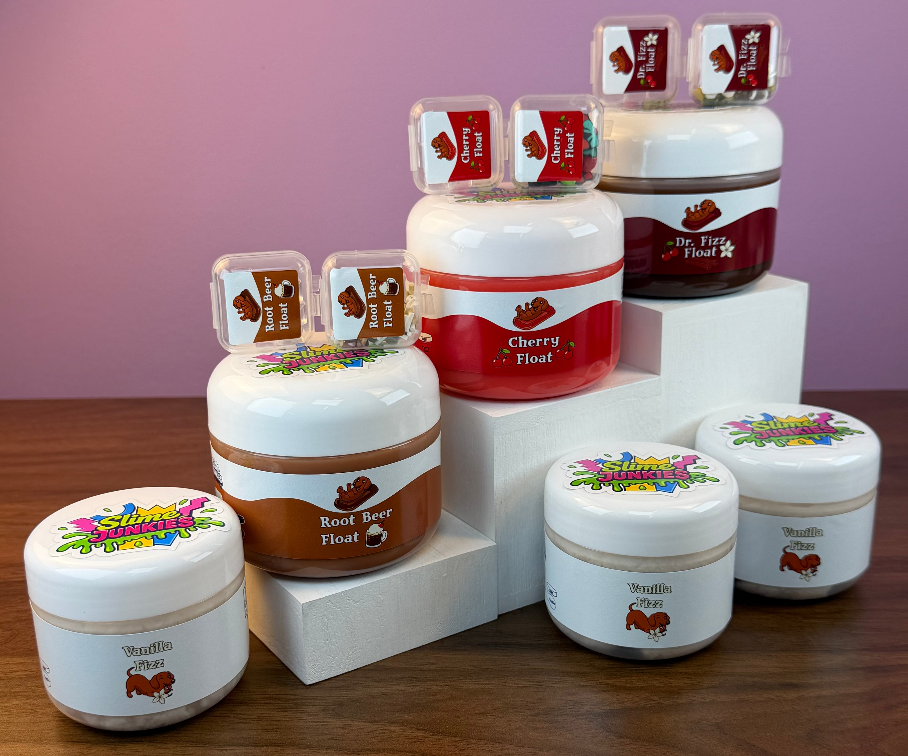 A set of slime jars with colorful labels, arranged on white display blocks over a wooden table against a pink background.