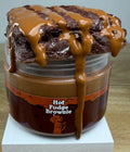 A novelty jar of 'Hot Fudge Brownie' slime, featuring a plastic jar with a brown label, topped with brown clay brownie with a brown slime drizzle, displayed on a white block.