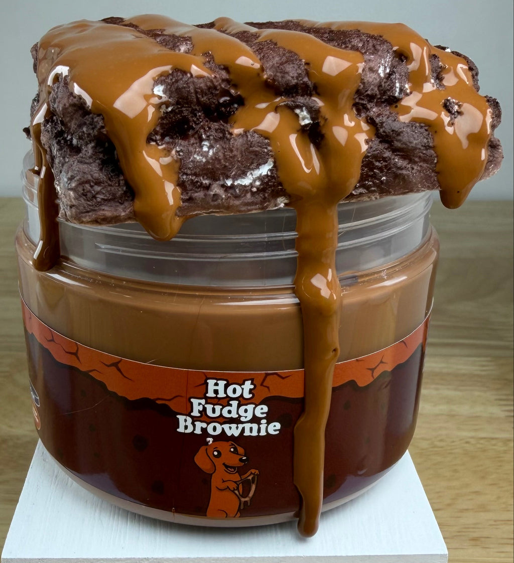 A novelty jar of 'Hot Fudge Brownie' slime, featuring a plastic jar with a brown label, topped with brown clay brownie with a brown slime drizzle, displayed on a white block.
