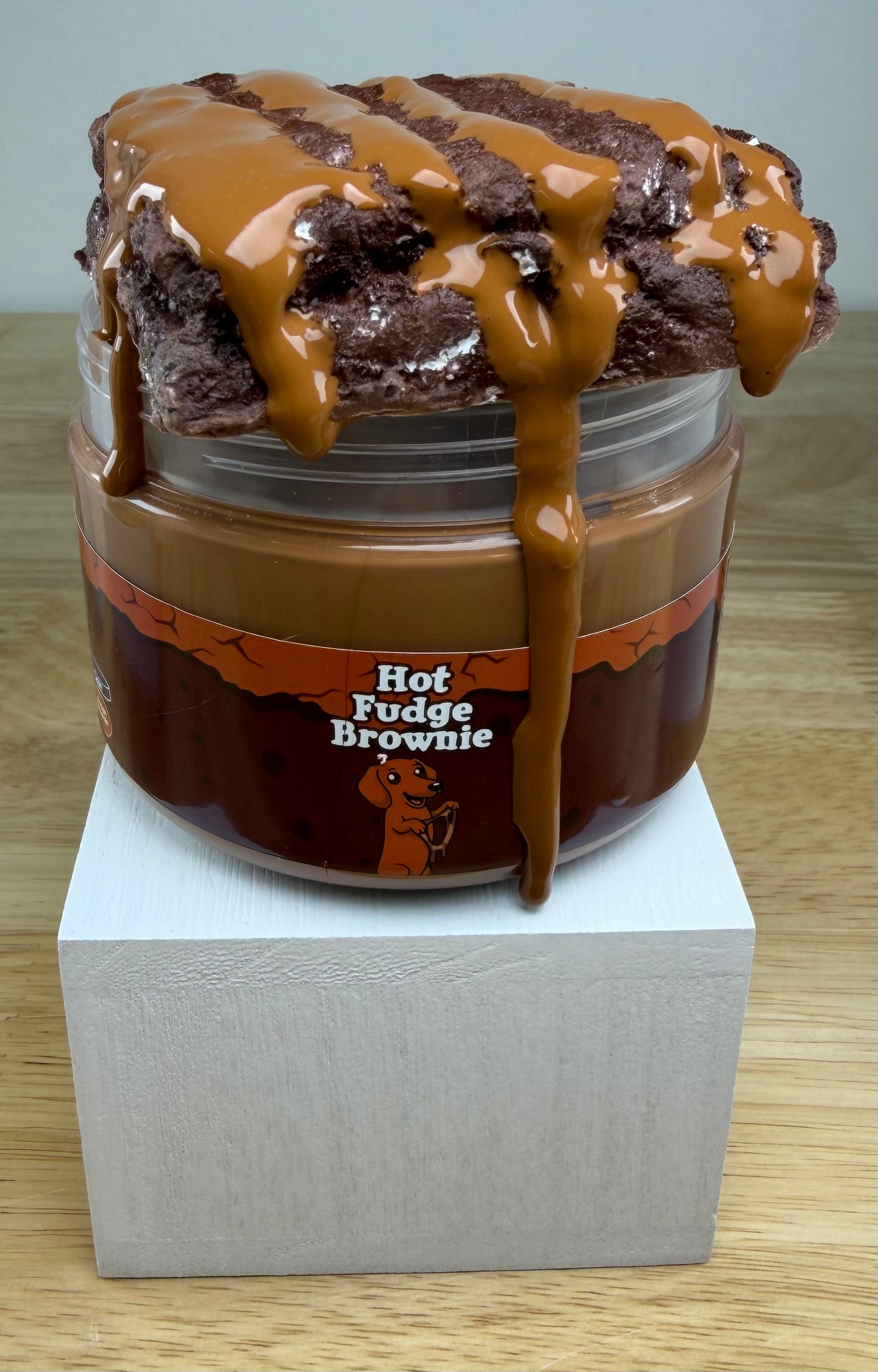A novelty jar of 'Hot Fudge Brownie' slime, featuring a plastic jar with a brown label, topped with brown clay brownie with a brown slime drizzle, displayed on a white block.