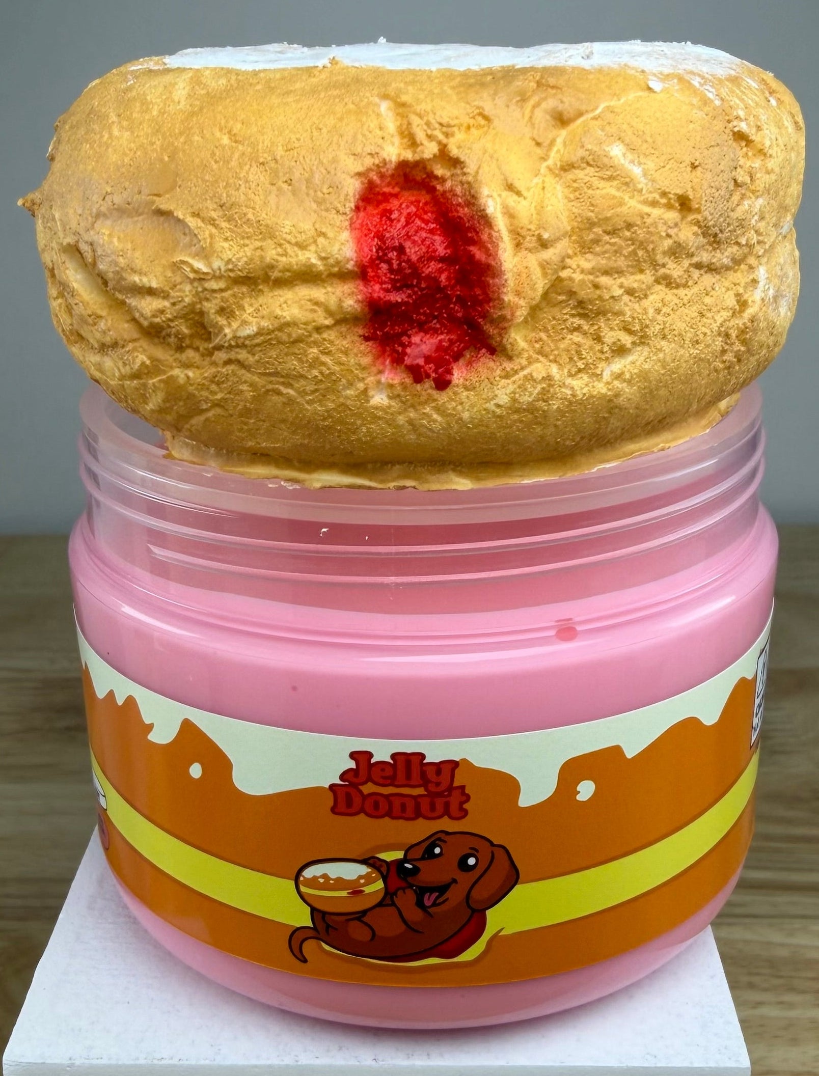 A novelty jar of 'Jelly Donut' slime or makeup, featuring a pink plastic jar with an orange and yellow label, topped with a textured golden-brown donut shape with a red center, displayed on a white block.