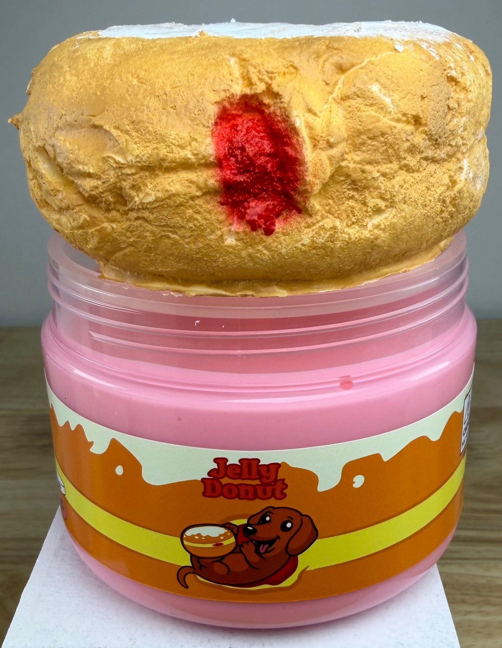 A novelty jar of 'Jelly Donut' slime or makeup, featuring a pink plastic jar with an orange and yellow label, topped with a textured golden-brown donut shape with a red center, displayed on a white block.
