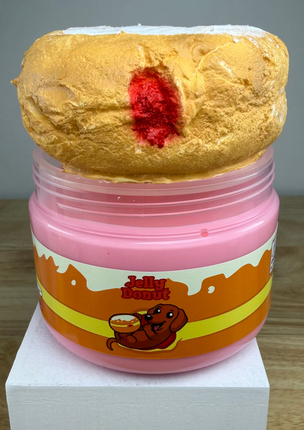 A novelty jar of 'Jelly Donut' slime or makeup, featuring a pink plastic jar with an orange and yellow label, topped with a textured golden-brown donut shape with a red center, displayed on a white block.
