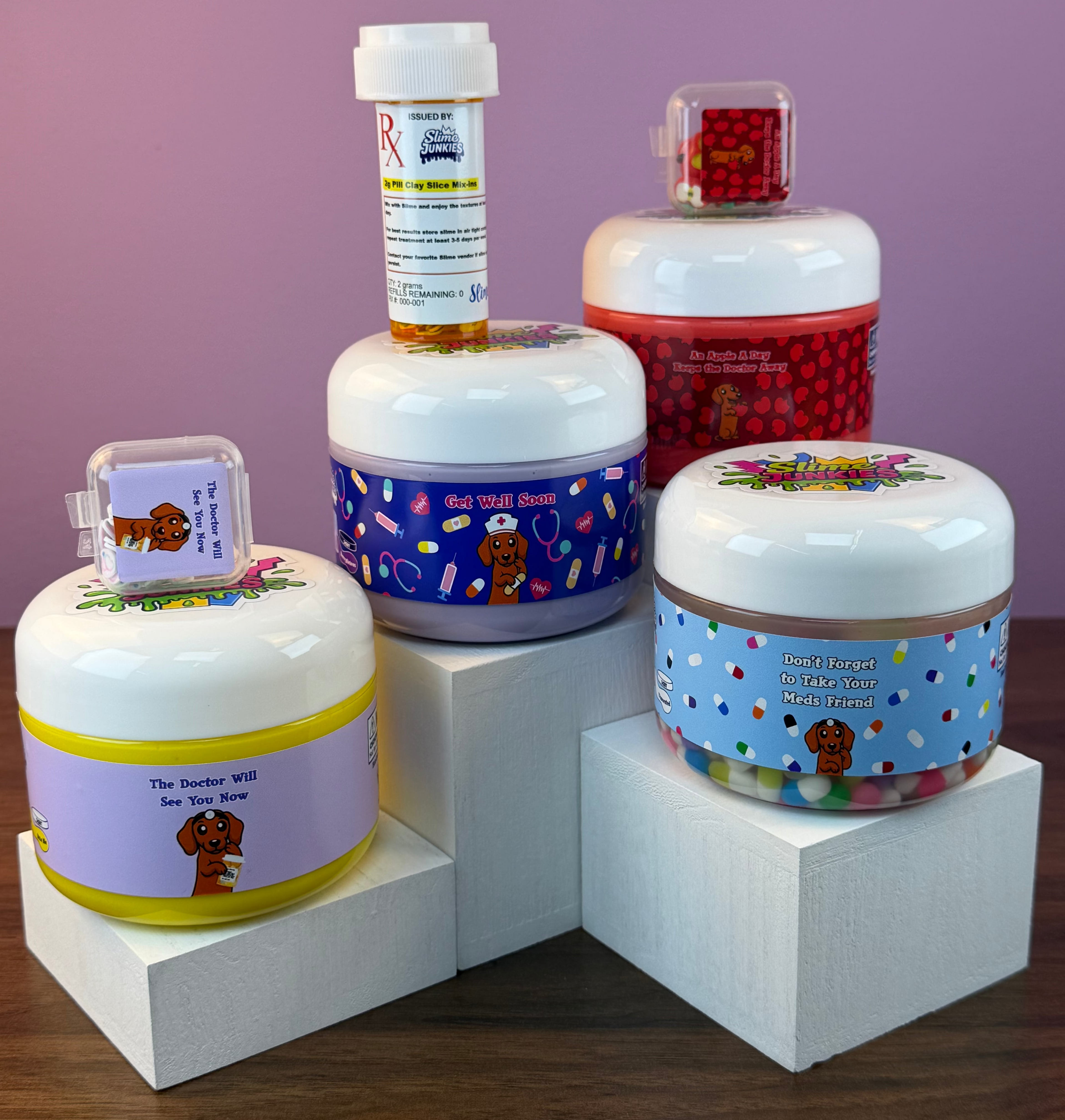 Four colorful slime jars and a pill bottle arranged on white display blocks against a lavender background. The slime jars have white lids and labels with dog illustrations, including one with a yellow band, one with a blue polka-dot pattern, and one with a red patterned band.