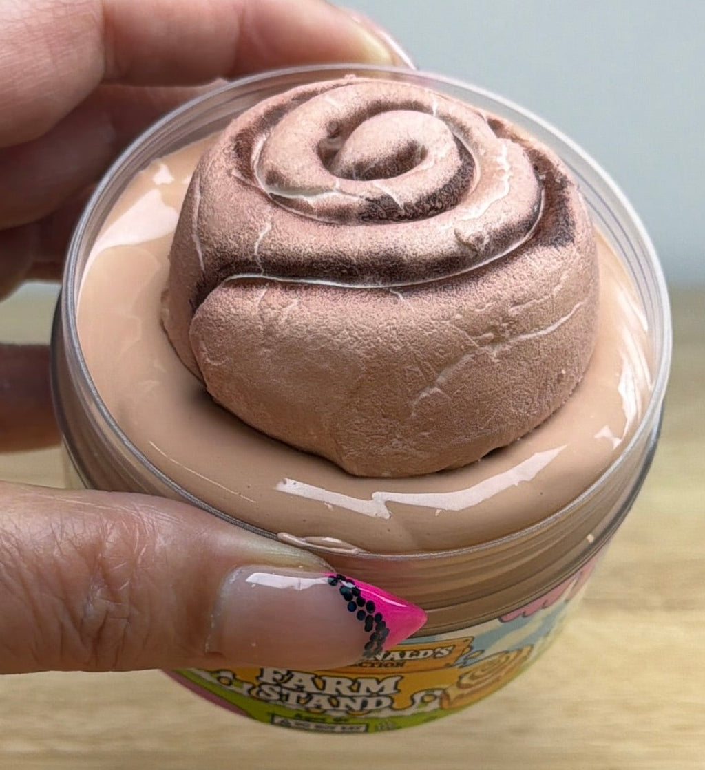 light tan slime with a clay cinnamon roll on it in a clear jar with an old macdonald farm collection farm label that says farm stand with a cinnamon roll on it