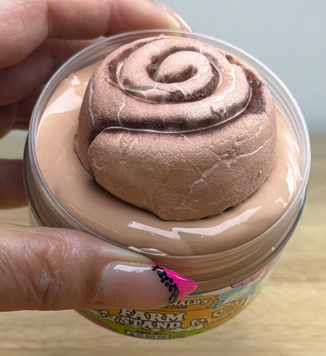 light tan slime with a clay cinnamon roll on it in a clear jar with an old macdonald farm collection farm label that says farm stand with a cinnamon roll on it