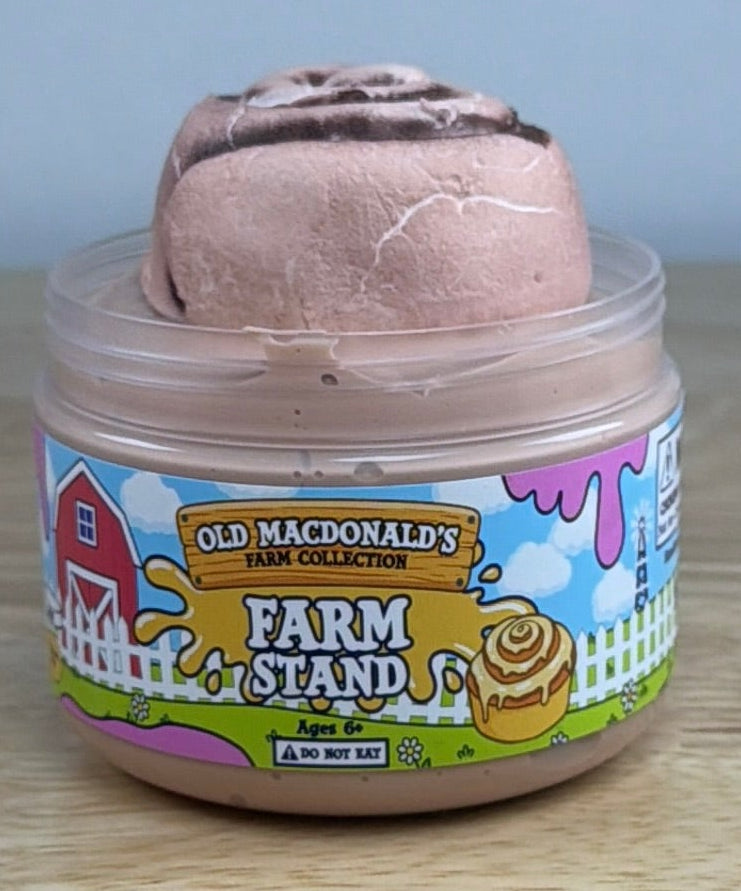 light tan slime with a clay cinnamon roll on it in a clear jar with an old macdonald farm collection farm label that says farm stand with a cinnamon roll on it