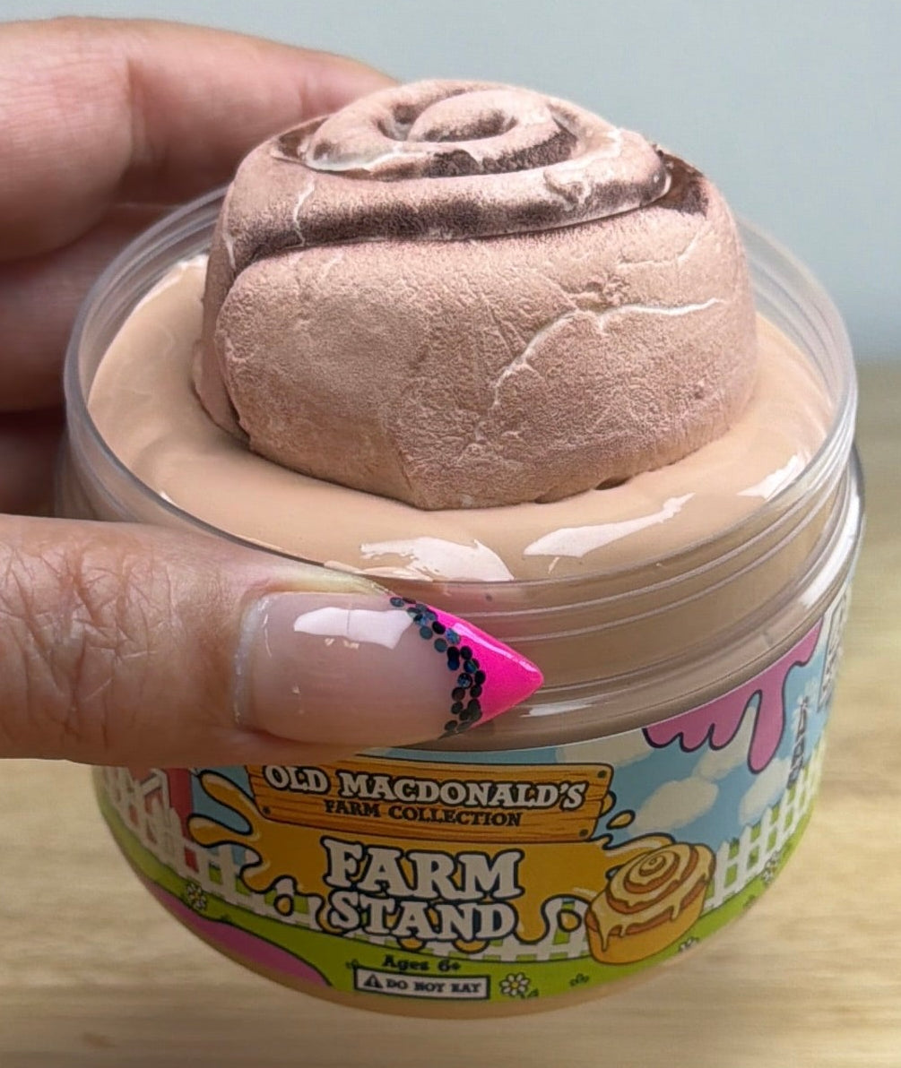 light tan slime with a clay cinnamon roll on it in a clear jar with an old macdonald farm collection farm label that says farm stand with a cinnamon roll on it