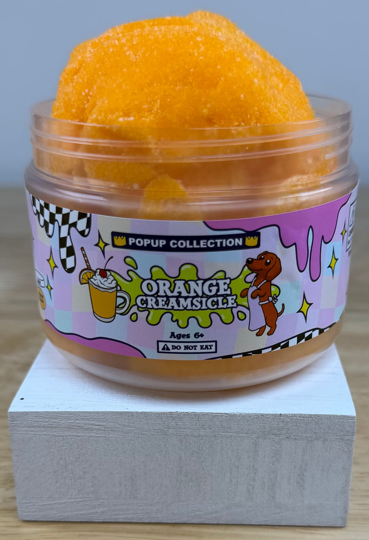 Orange cloud slime in a pop up collection labelled plastic jar that says orange creamsicle with a dog and a cup with orange dessert in it.