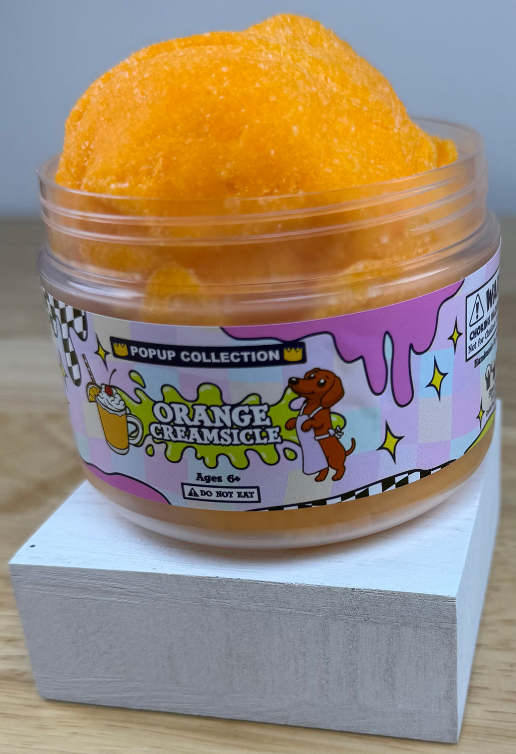 Orange cloud slime in a pop up collection labelled plastic jar that says orange creamsicle with a dog and a cup with orange dessert in it.