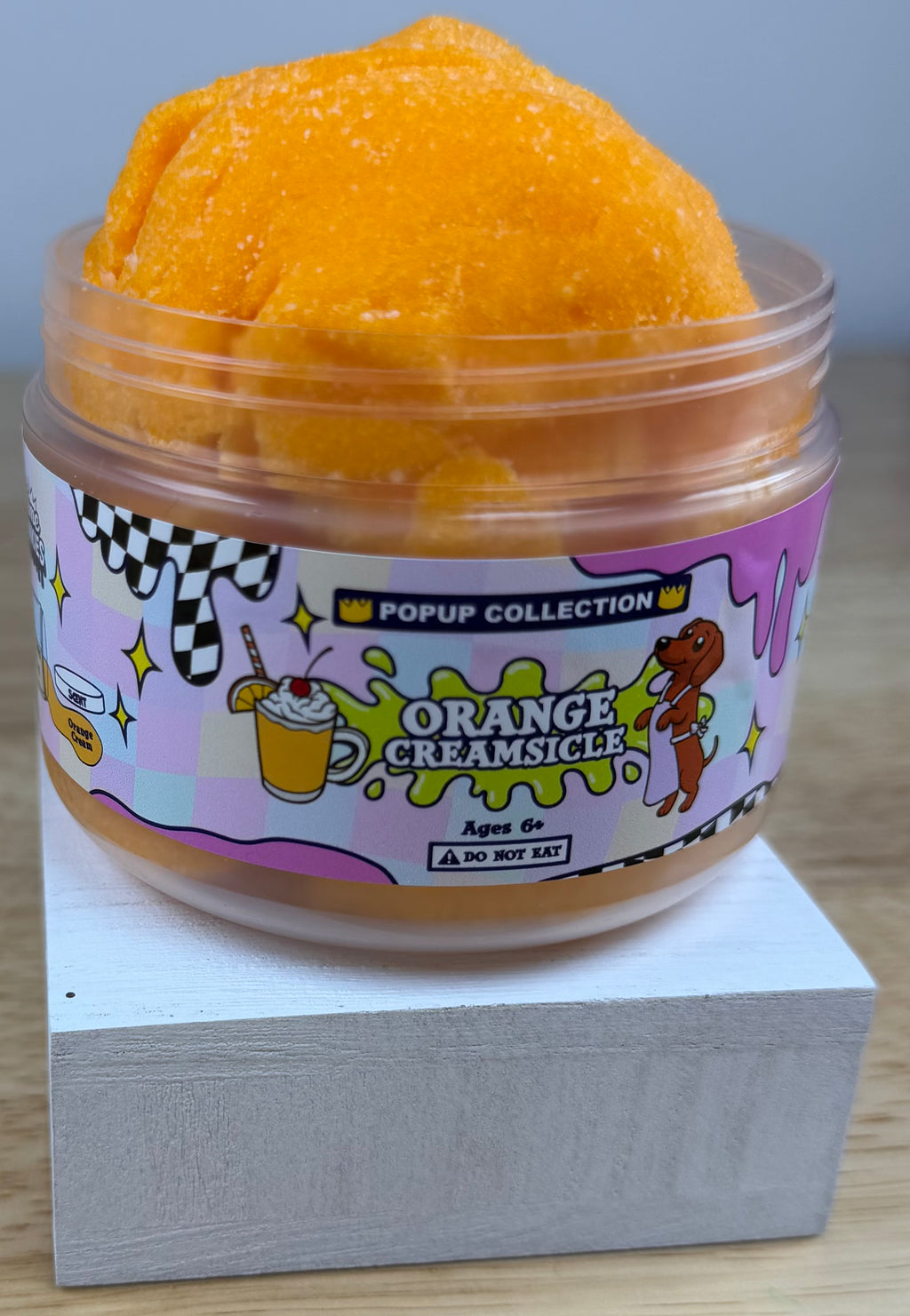 Orange cloud slime in a pop up collection labelled plastic jar that says orange creamsicle with a dog and a cup with orange dessert in it.