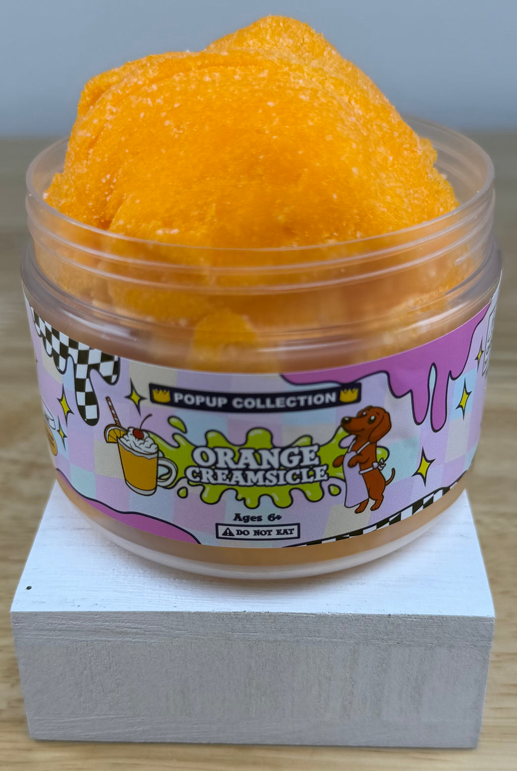 Orange cloud slime in a pop up collection labelled plastic jar that says orange creamsicle with a dog and a cup with orange dessert in it.