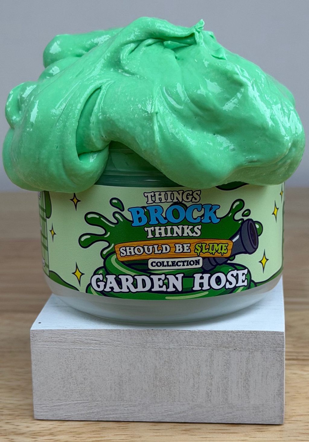 A jar of green slime with a matching green slime mound on top, displayed on a white block over a wooden surface against a white background. The label reads 'Things Brock Thinks Should Be Slime Collector's Edition Garden Hose'.