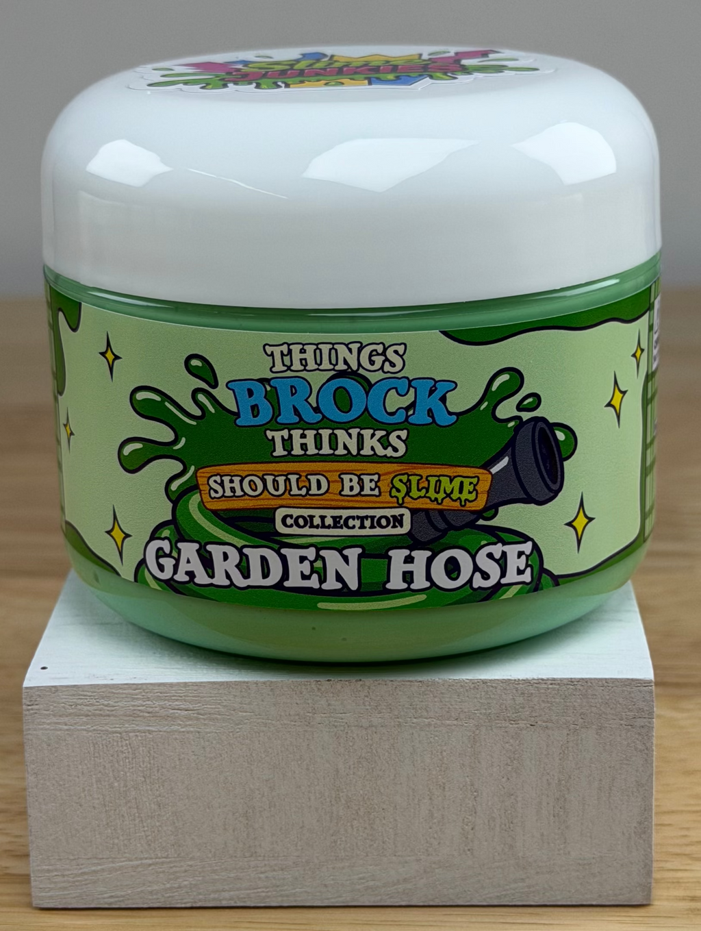 A jar of green slime with a matching green slime mound on top, displayed on a white block over a wooden surface against a white background. The label reads 'Things Brock Thinks Should Be Slime Collector's Edition Garden Hose'.