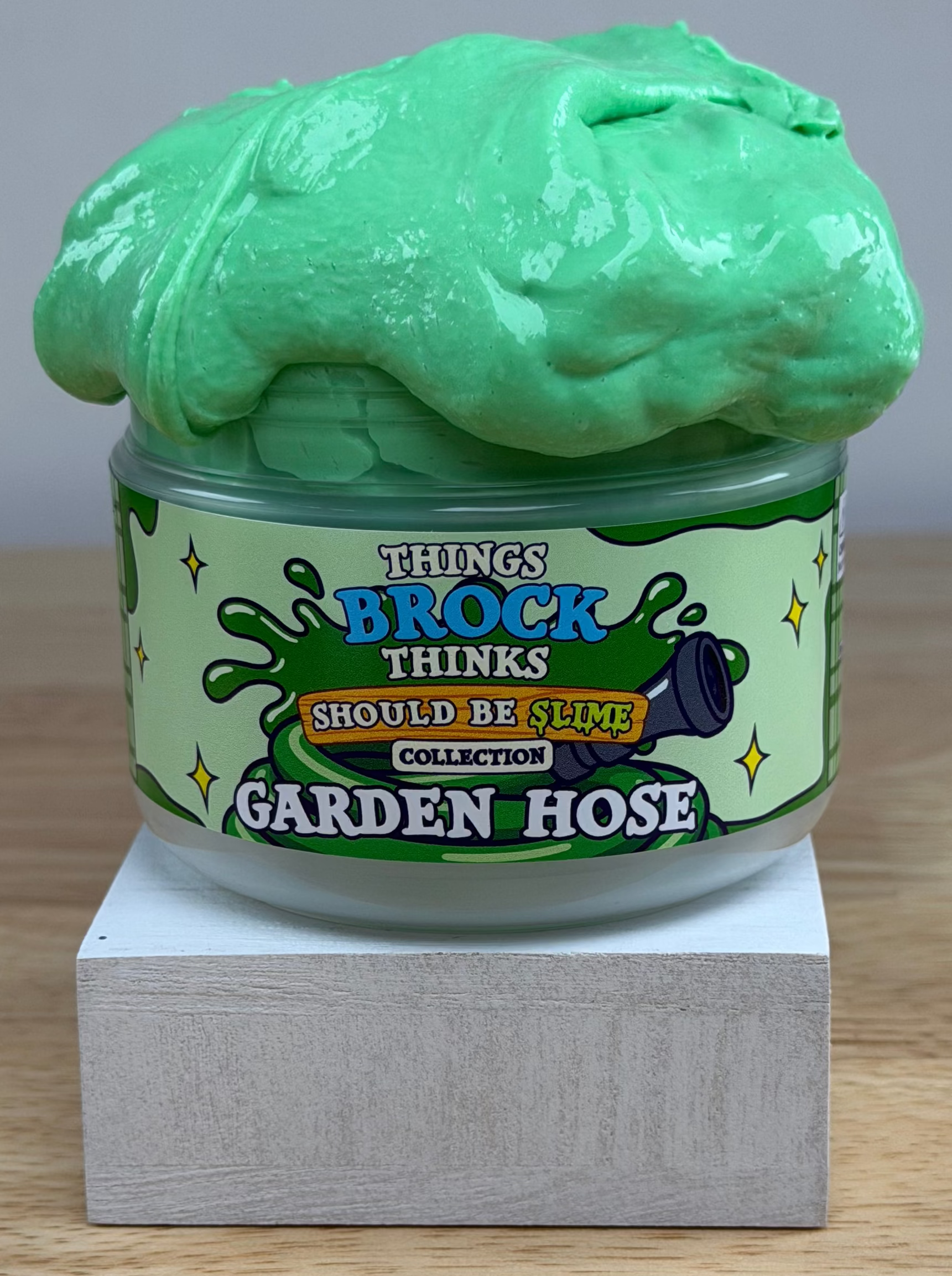 A jar of green slime with a matching green slime mound on top, displayed on a white block over a wooden surface against a white background. The label reads 'Things Brock Thinks Should Be Slime Collector's Edition Garden Hose'.