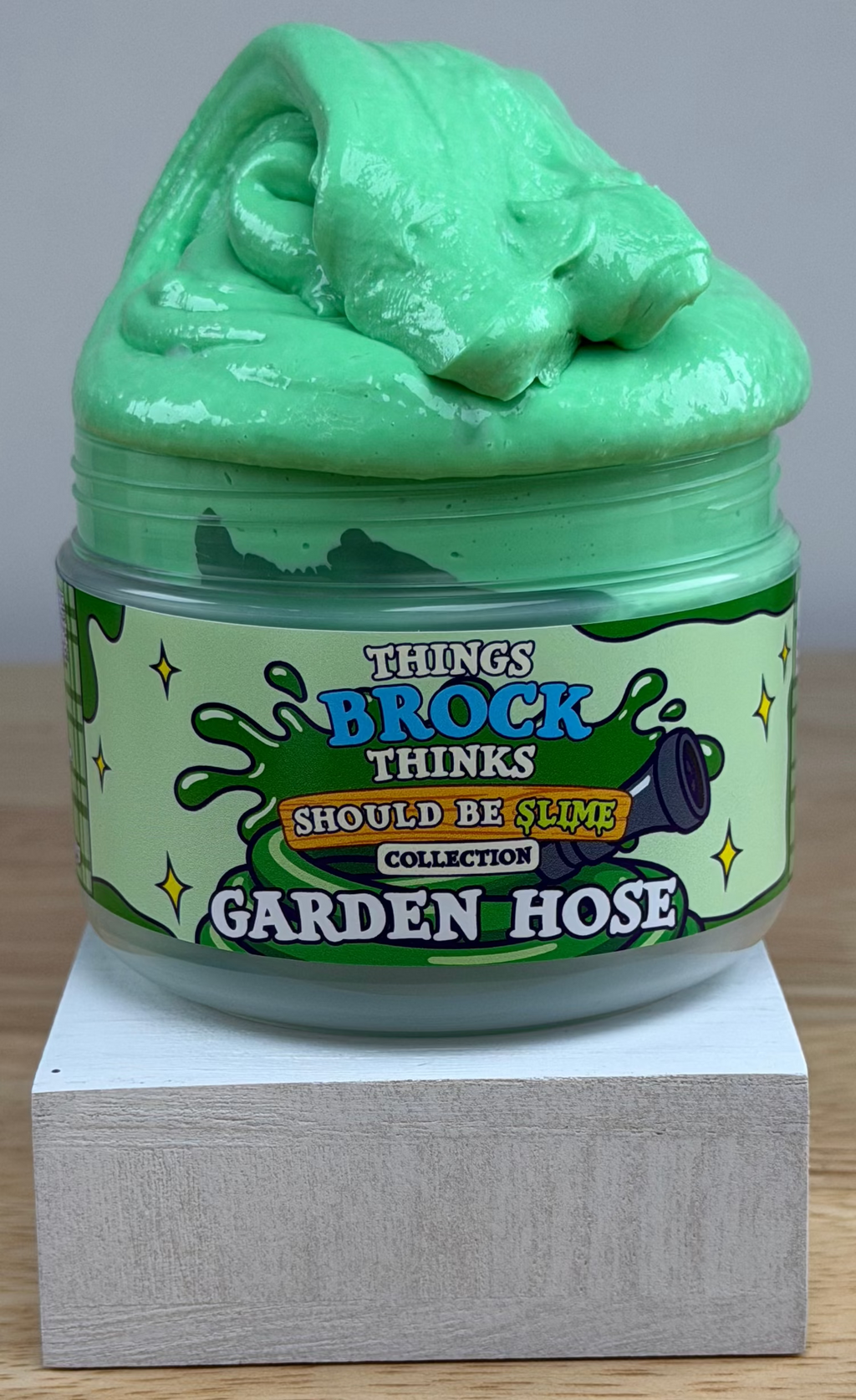 A jar of green slime with a matching green slime mound on top, displayed on a white block over a wooden surface against a white background. The label reads 'Things Brock Thinks Should Be Slime Collector's Edition Garden Hose'.