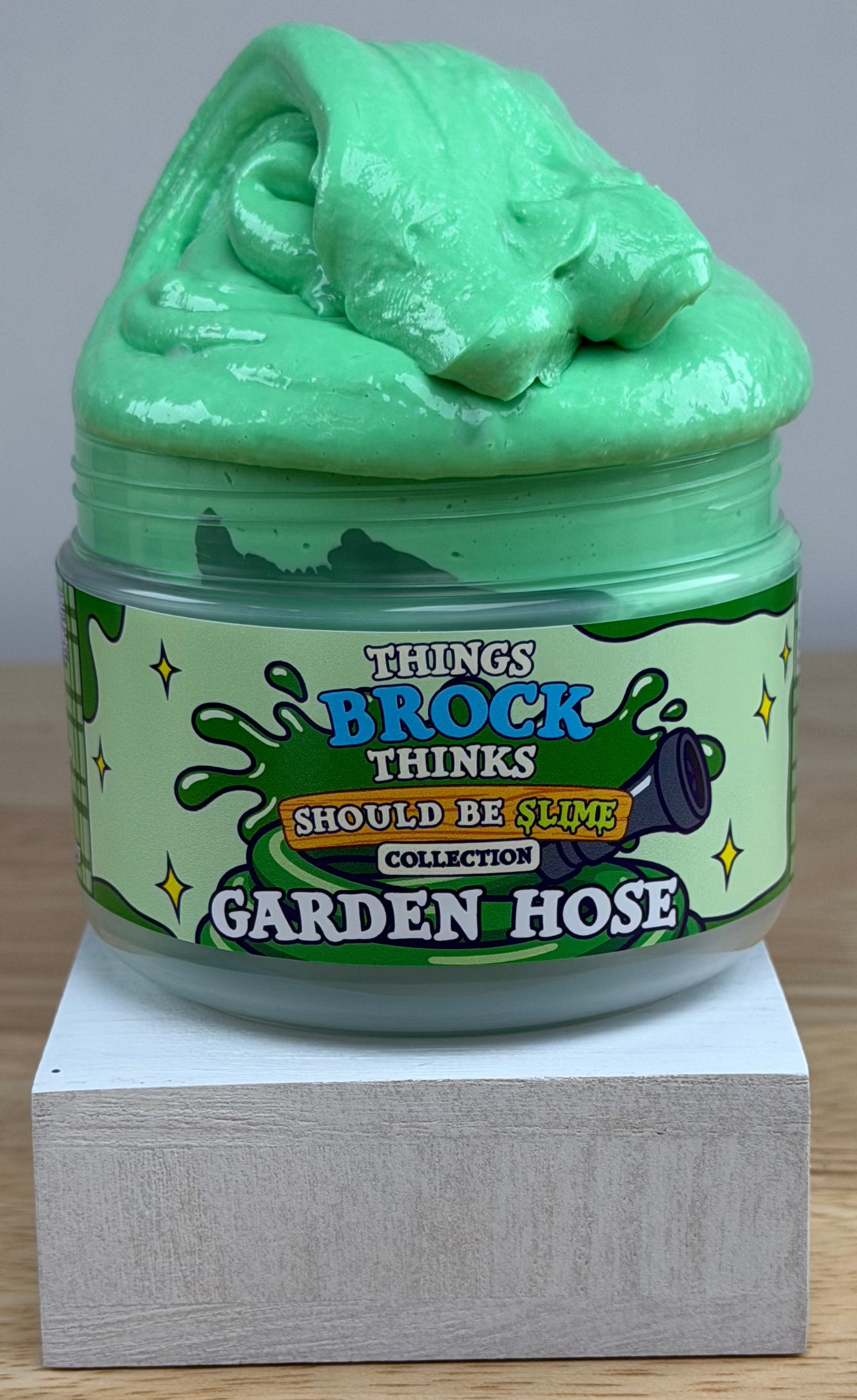 A jar of green slime with a matching green slime mound on top, displayed on a white block over a wooden surface against a white background. The label reads 'Things Brock Thinks Should Be Slime Collector's Edition Garden Hose'.