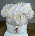 A tub of pastel slime with soft pink, lavender, and white tones, topped with multicolored foam beads, in a clear plastic container labeled 'Slime N Dots' on a white display block.