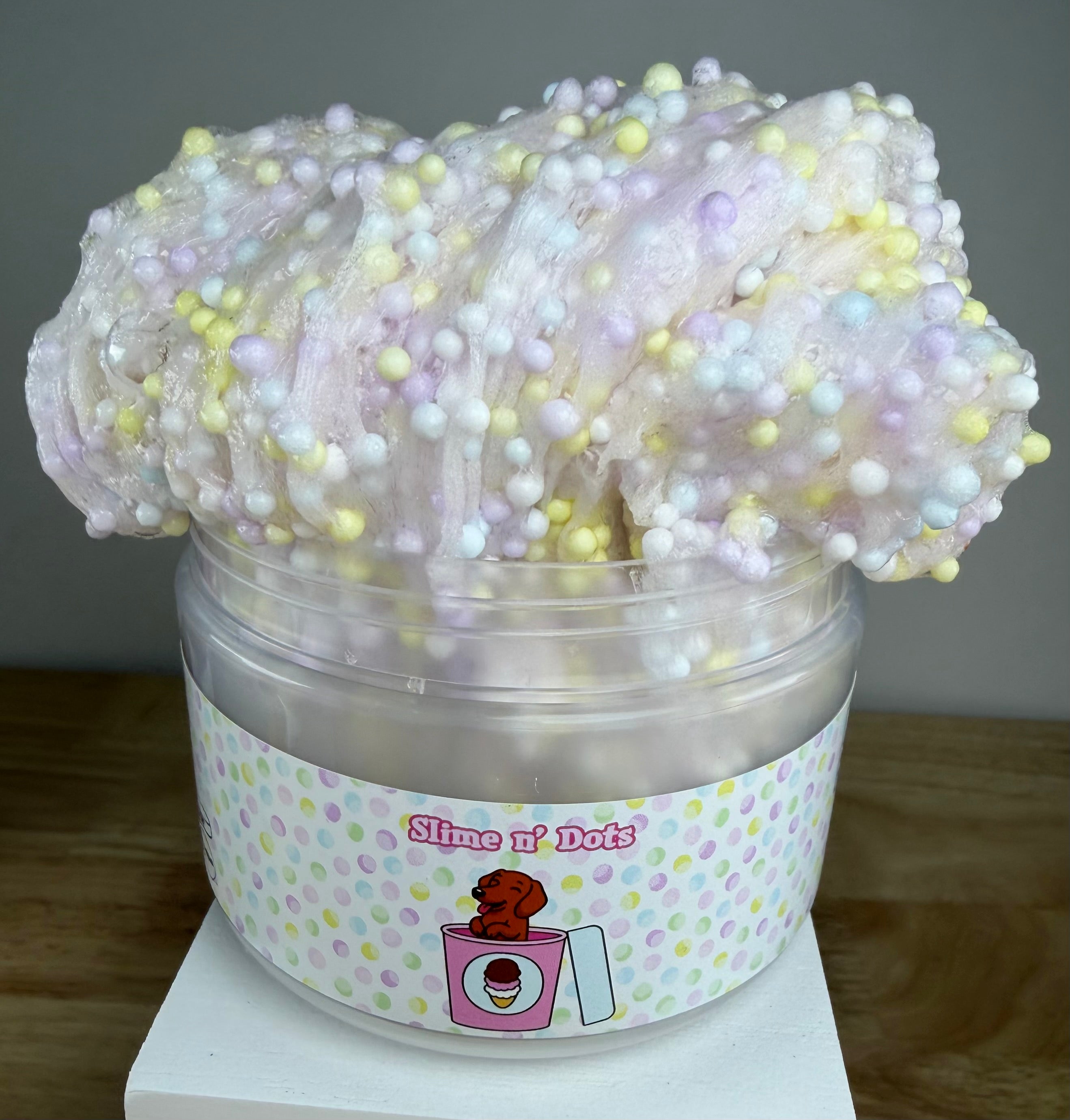 A tub of pastel slime with soft pink, lavender, and white tones, topped with multicolored foam beads, in a clear plastic container labeled 'Slime N Dots' on a white display block.