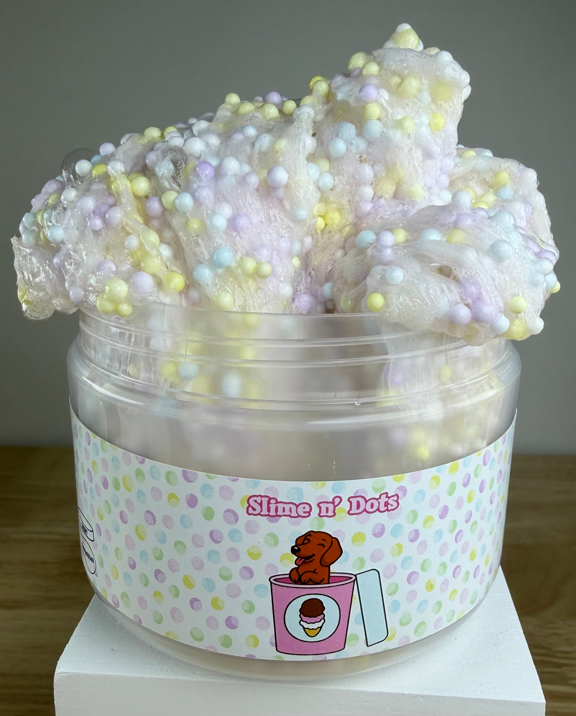 A tub of pastel slime with soft pink, lavender, and white tones, topped with multicolored foam beads, in a clear plastic container labeled 'Slime N Dots' on a white display block.