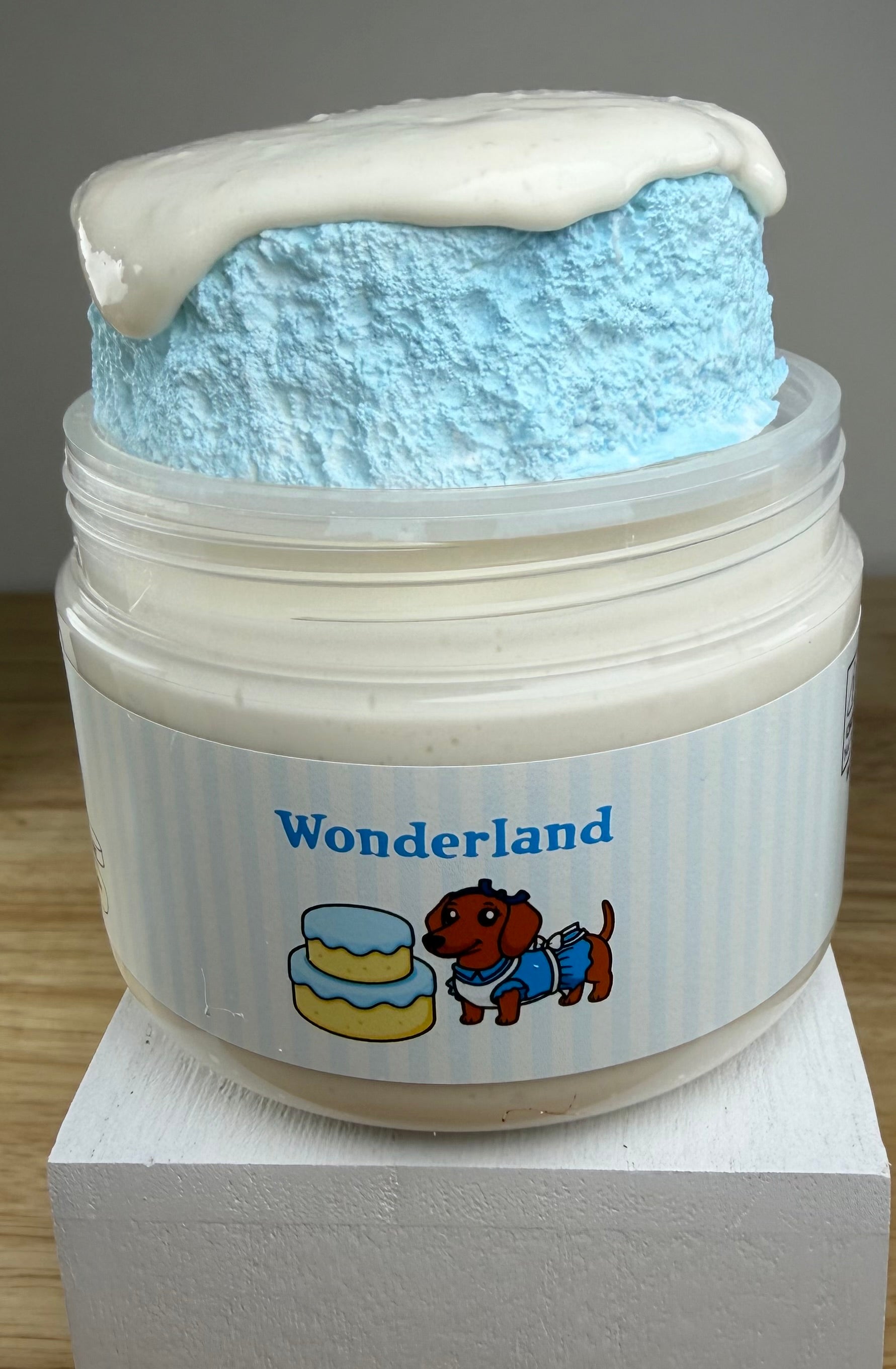 A novelty jar of 'Wonderland' slime, featuring a plastic jar with a white and light blue label, topped with light blue clay cake with a white slime drizzle, displayed on a white block.