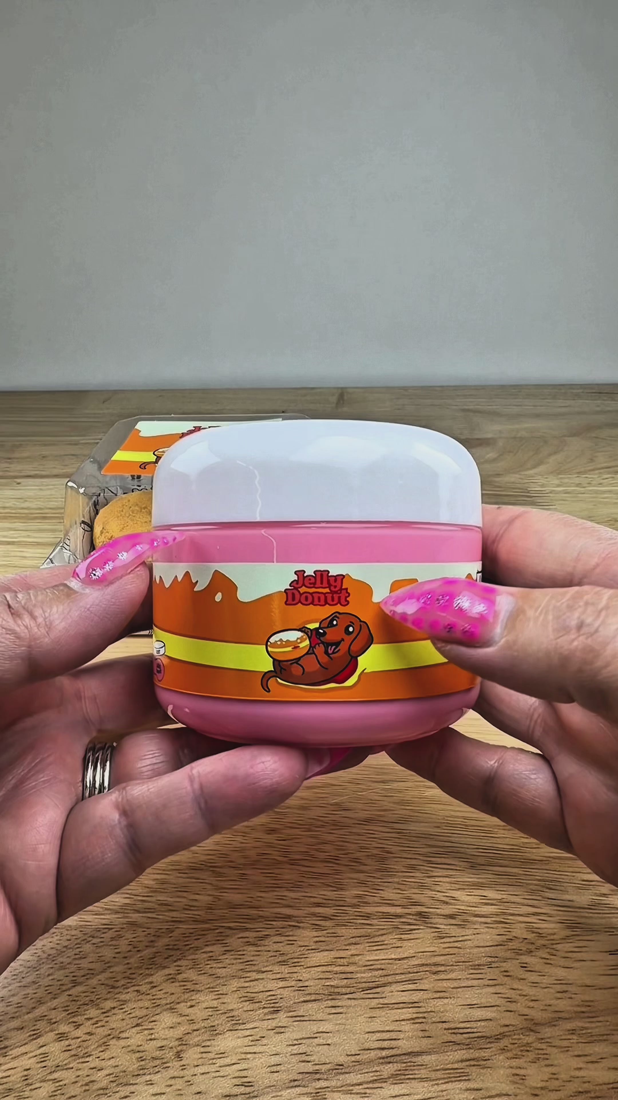 A novelty jar of 'Jelly Donut' slime or makeup, featuring a pink plastic jar with an orange and yellow label, topped with a textured golden-brown donut shape with a red center, displayed on a white block.