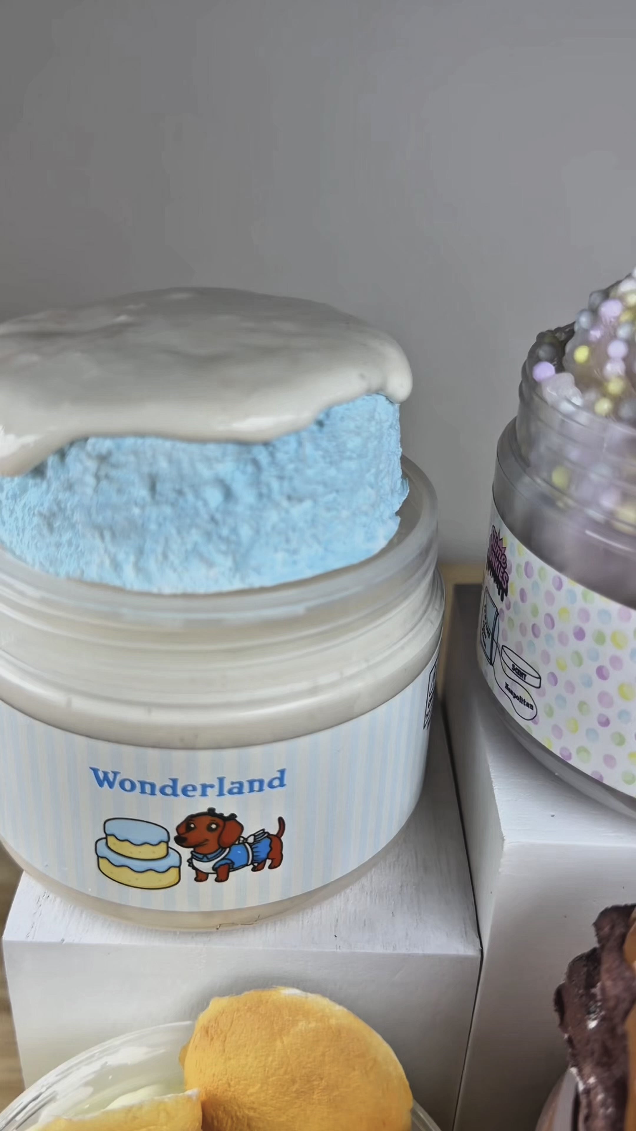 Five jars of squishy slime displayed on white display blocks on a wooden surface. The jars feature creative, food-inspired slimes with clay toppers resembling donuts, cakes, bananas, cookie wafers, and brownies with a chocolate slime drizzle.