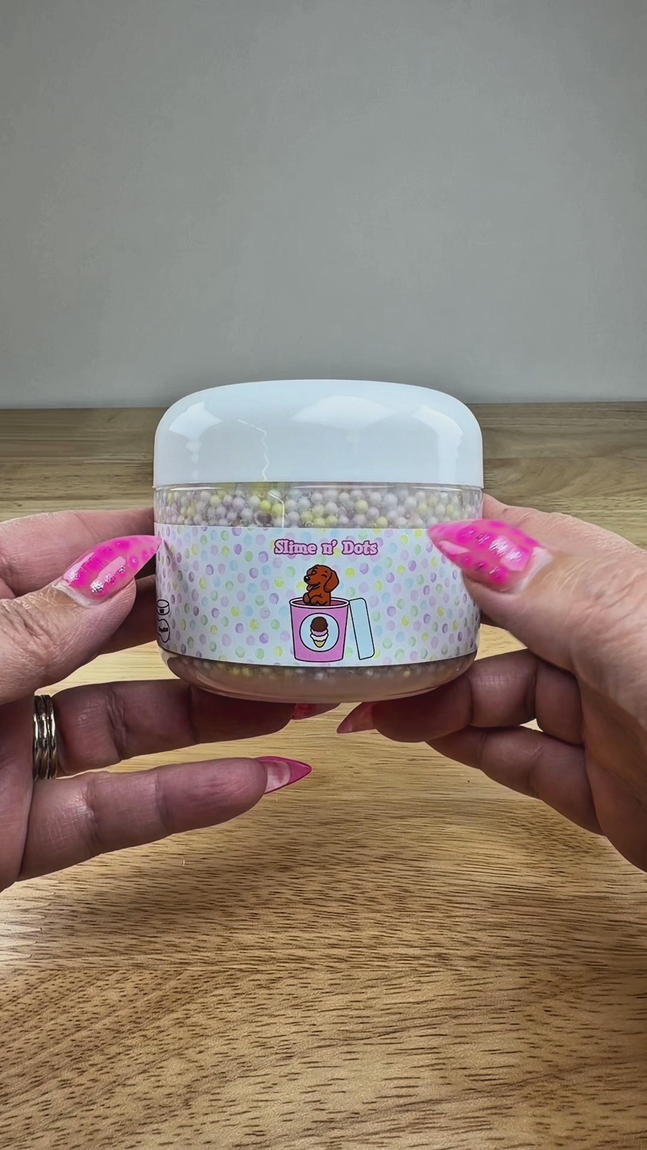 A tub of pastel slime with soft pink, lavender, and white tones, topped with multicolored foam beads, in a clear plastic container labeled 'Slime N Dots' on a white display block.