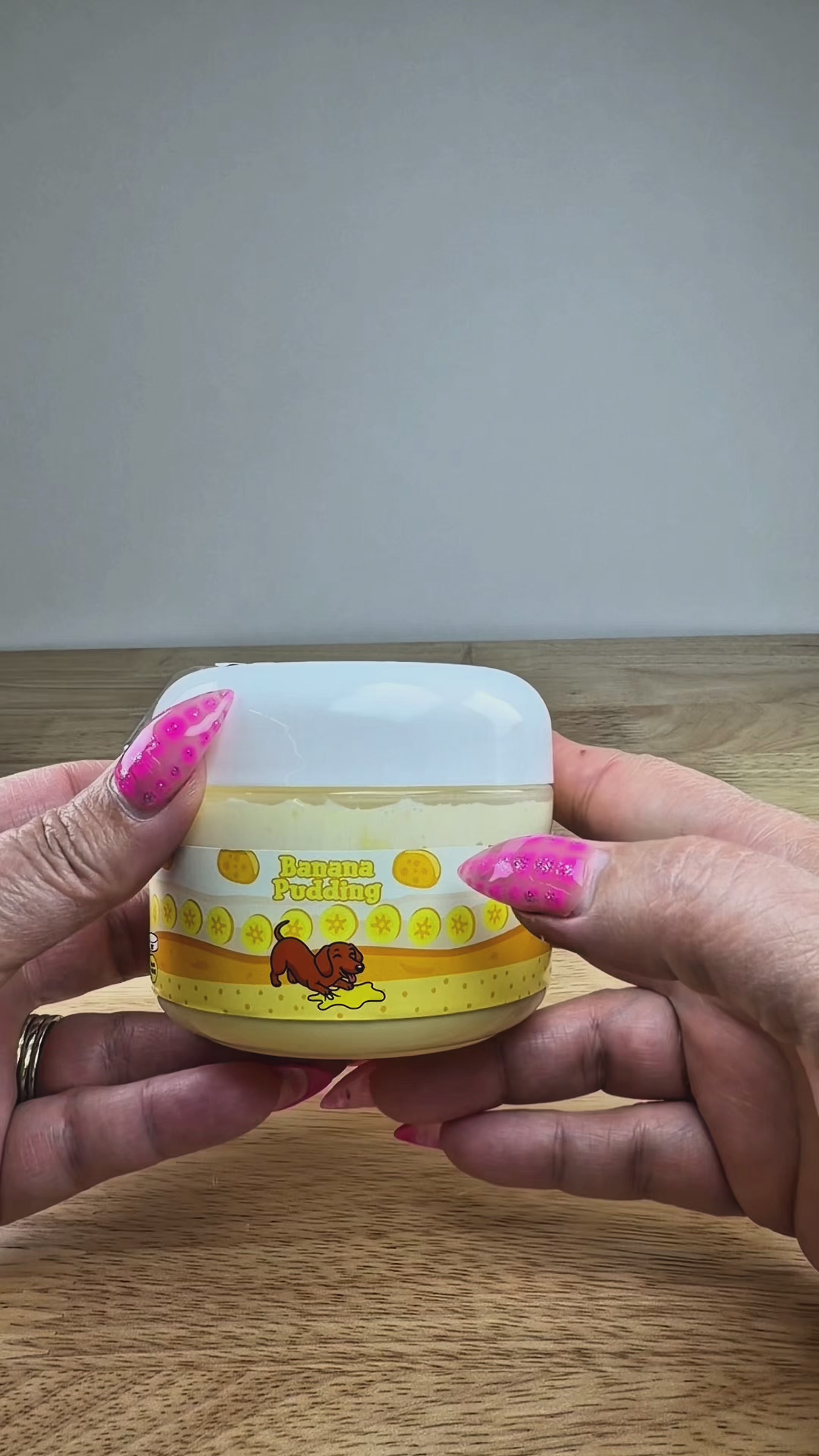 A novelty jar of 'Banana Pudding' slime, featuring a plastic jar with a yellow and orange label, topped with yellow clay banana slices with light tan clay cookie wafers, displayed on a white block.